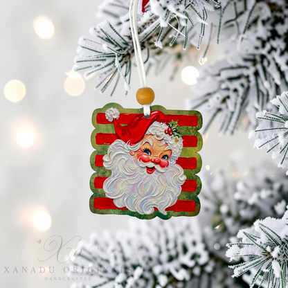 A square Christmas ornament featuring a vintage-style Santa Claus face with red and green striped background, hanging from a white string on a frosted Christmas tree branch with warm bokeh lights in the background.