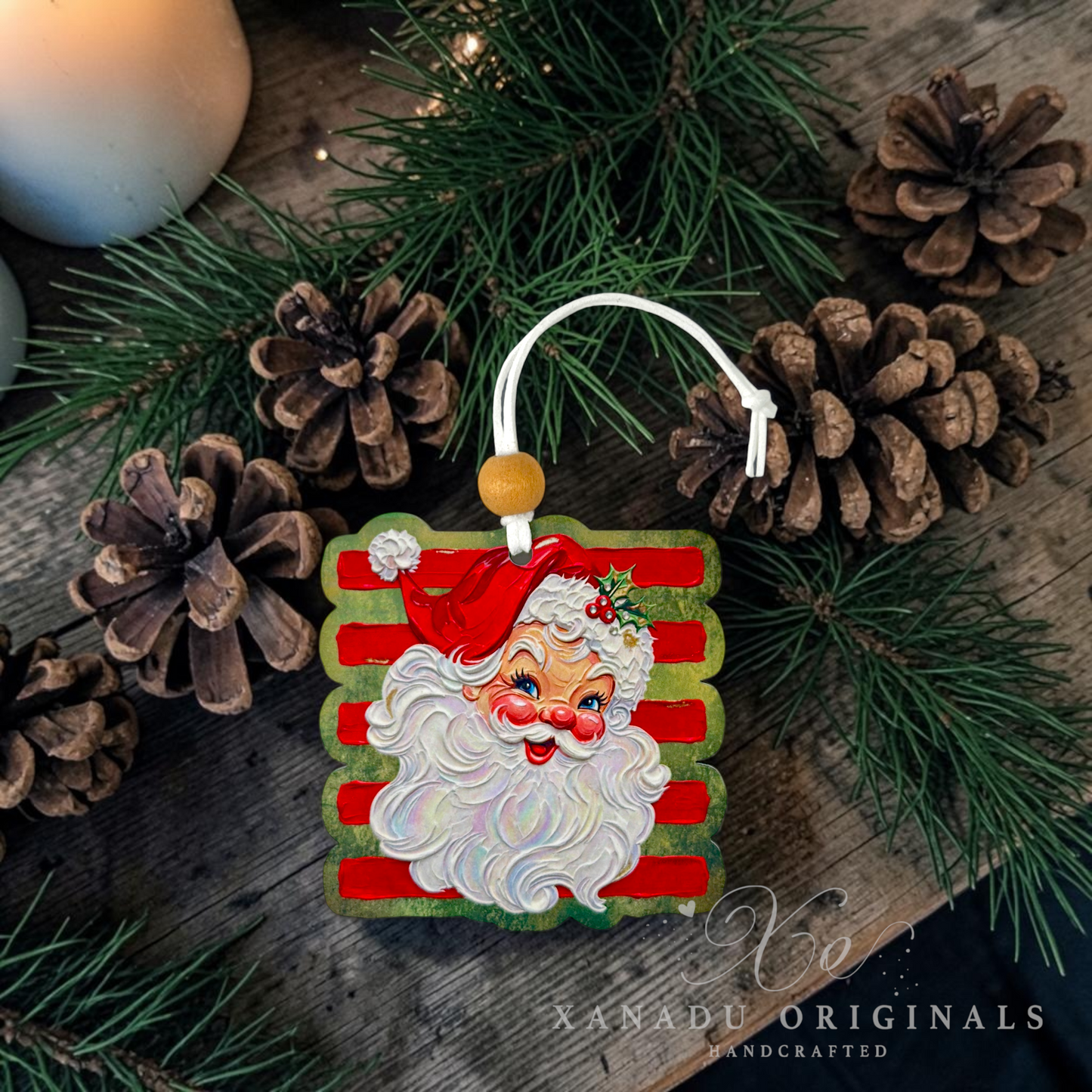 A square Christmas ornament featuring a painted Santa Claus face with red and white striped background, hanging from a white cord with a wooden bead. The ornament is styled on a rustic wooden surface with pine branches, pinecones, and a candle, with the watermark 'Xanadu Originals Handcrafted' in the lower right.