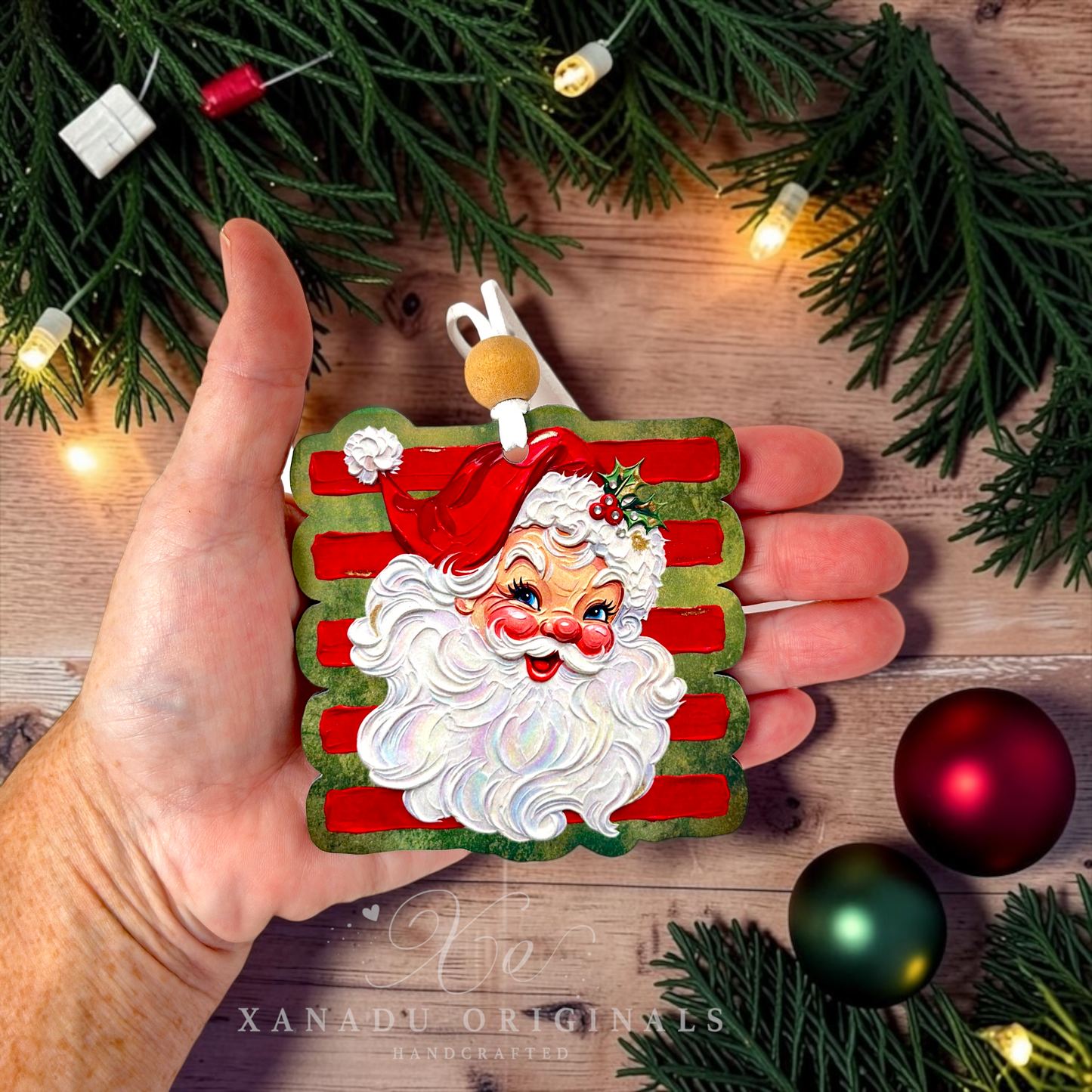 A hand holds a square Christmas ornament featuring a smiling Santa face with a white beard, red hat, and red-and-white striped background. The ornament is photographed on a rustic wooden surface with evergreen branches, warm string lights, and holiday ornaments around it.
