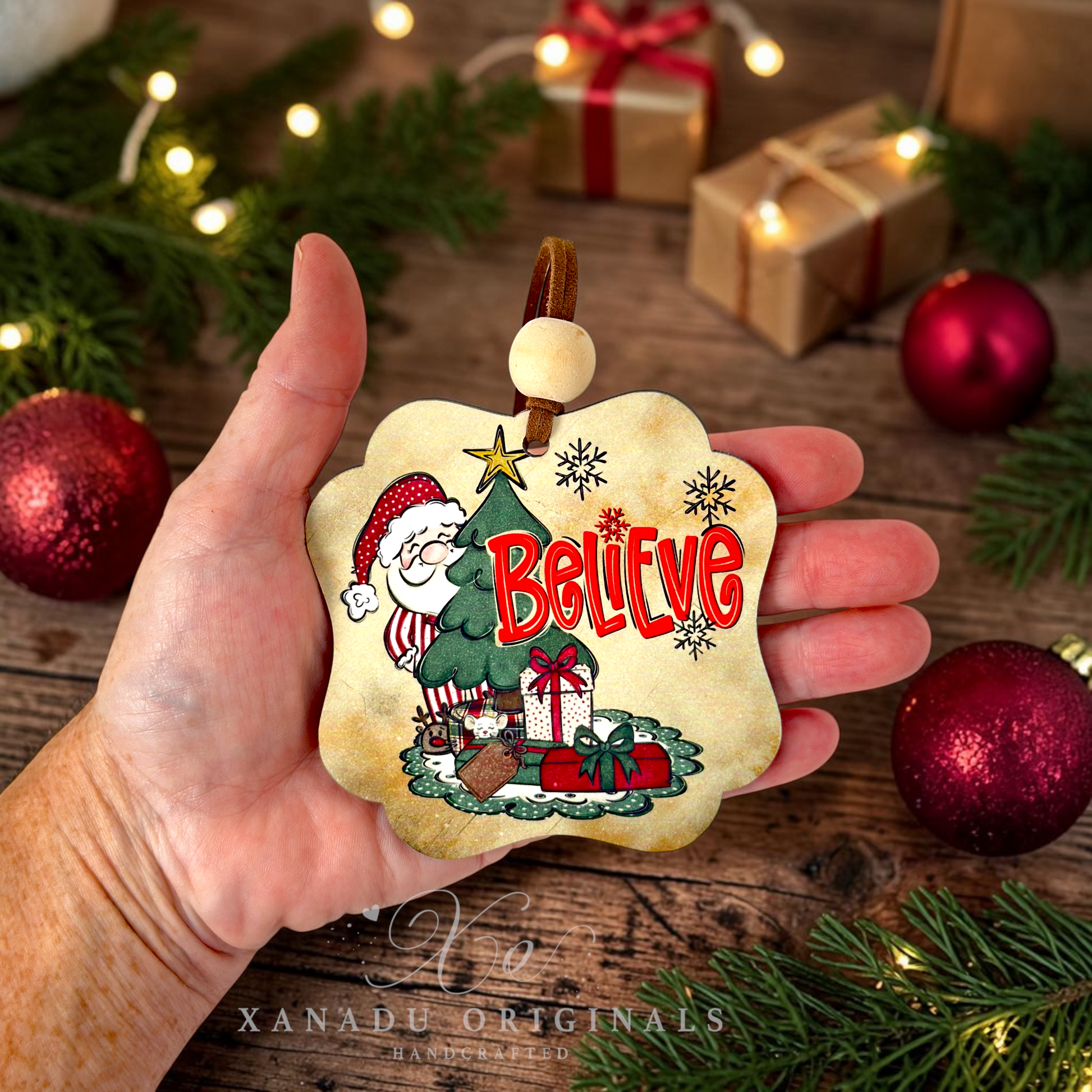 A hand holds a decorative Christmas ornament featuring an illustrated Santa Claus, a Christmas tree, gifts, and the word “Believe” in red. The scene is styled on a rustic wooden surface with pine branches, string lights, wrapped presents, and red ornaments.