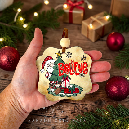 A hand holds a decorative Christmas ornament featuring an illustrated Santa Claus, a Christmas tree, gifts, and the word “Believe” in red. The scene is styled on a rustic wooden surface with pine branches, string lights, wrapped presents, and red ornaments.