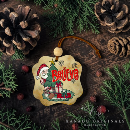 A scalloped Christmas ornament featuring a cartoon Santa Claus by a tree with the word 'Believe' in red, surrounded by pinecones, evergreen branches, and small red berries on a dark wood surface. A watermark for 'XANADU ORIGINALS' is visible at the bottom.