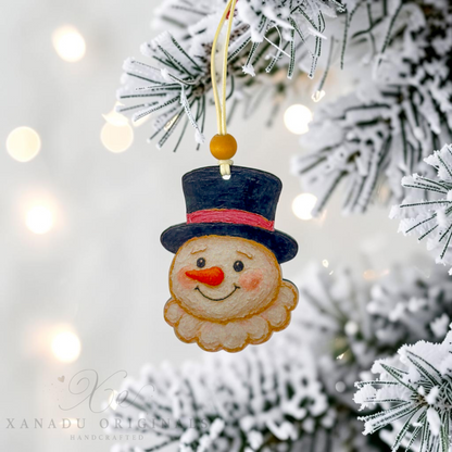 A handcrafted felt Christmas snowman ornament hanging from a flocked Christmas tree branch with warm bokeh lights in the background. The ornament features a beige felt face with a black top hat, red band, red nose, and pink cheeks, with a gold hanging string.