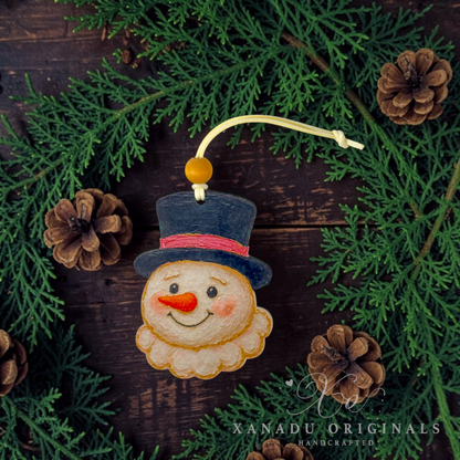 A handmade felt snowman ornament with a dark blue top hat, pink band, orange carrot nose, and cream beard, hanging from a yellow cord. The ornament is styled on a dark wooden surface surrounded by green pine branches and pinecones.