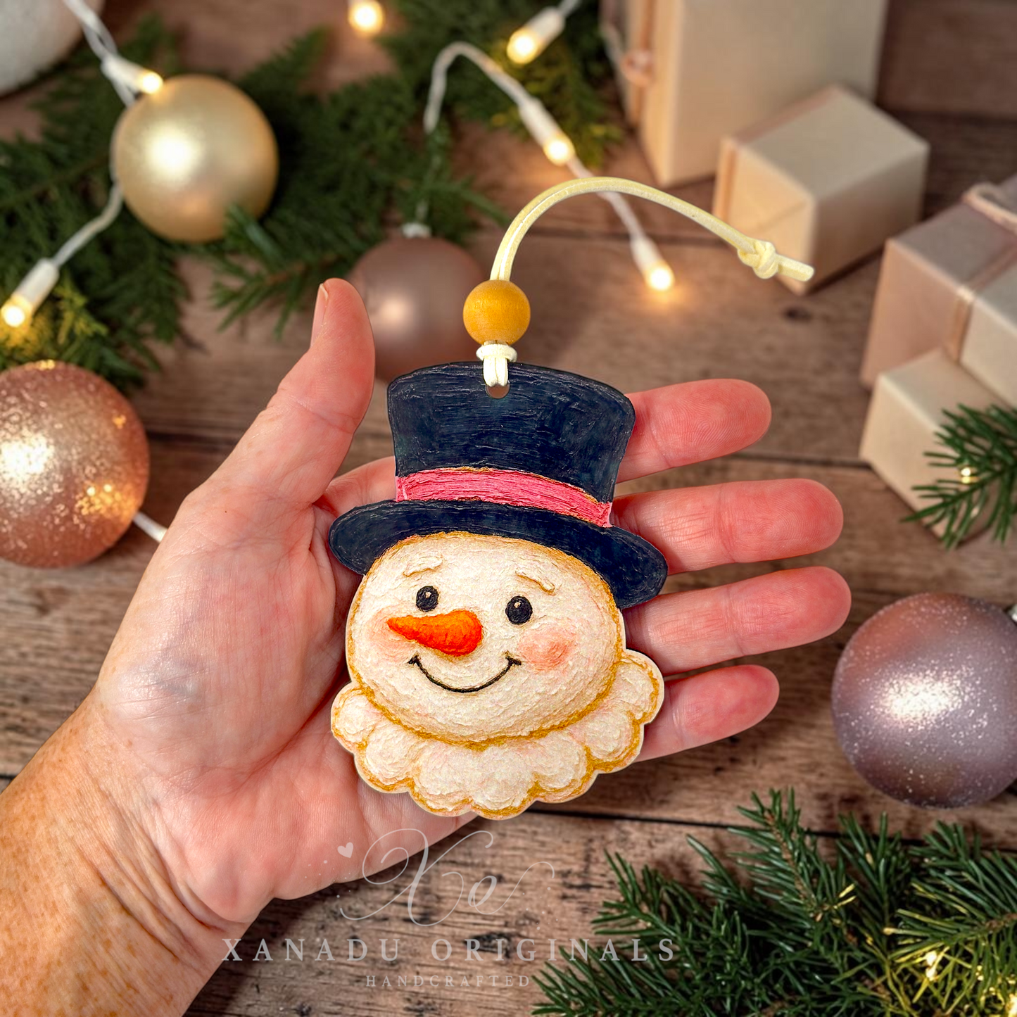A hand holds a handmade felt snowman ornament with a black top hat, pink band, orange carrot nose, and cream beard. The background shows a rustic wooden table with Christmas ornaments, pine branches, string lights, and gift boxes.