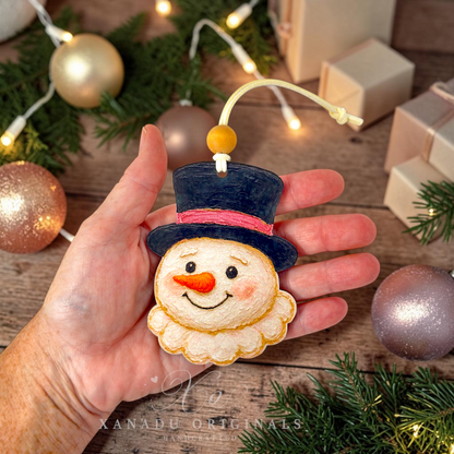 A hand holds a handmade felt snowman ornament with a black top hat, pink band, orange carrot nose, and cream beard. The background shows a rustic wooden table with Christmas ornaments, pine branches, string lights, and gift boxes.