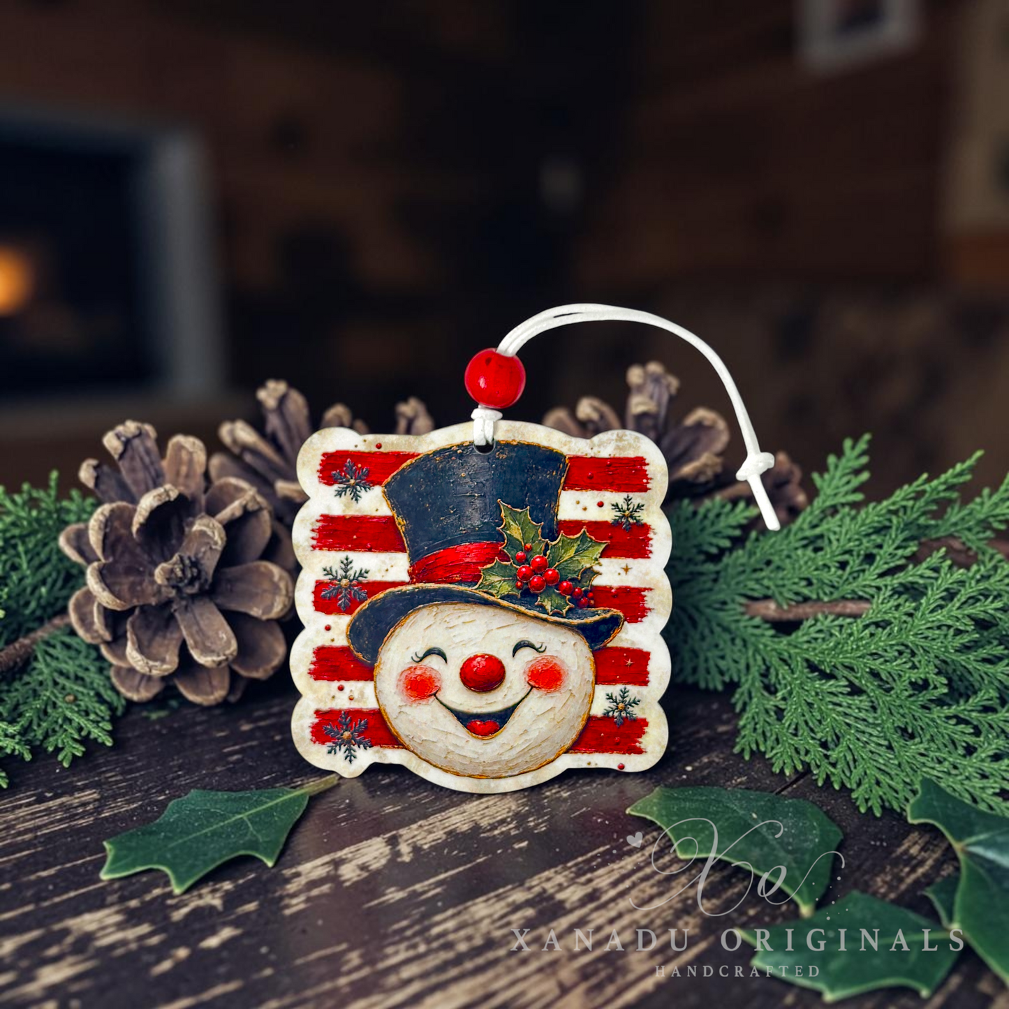 A handcrafted wooden Christmas ornament shaped like a smiling snowman face with a red striped hat, displayed on a rustic wooden surface with pinecones, evergreen branches, and holly leaves. The ornament has a white hanging loop with a red bead.