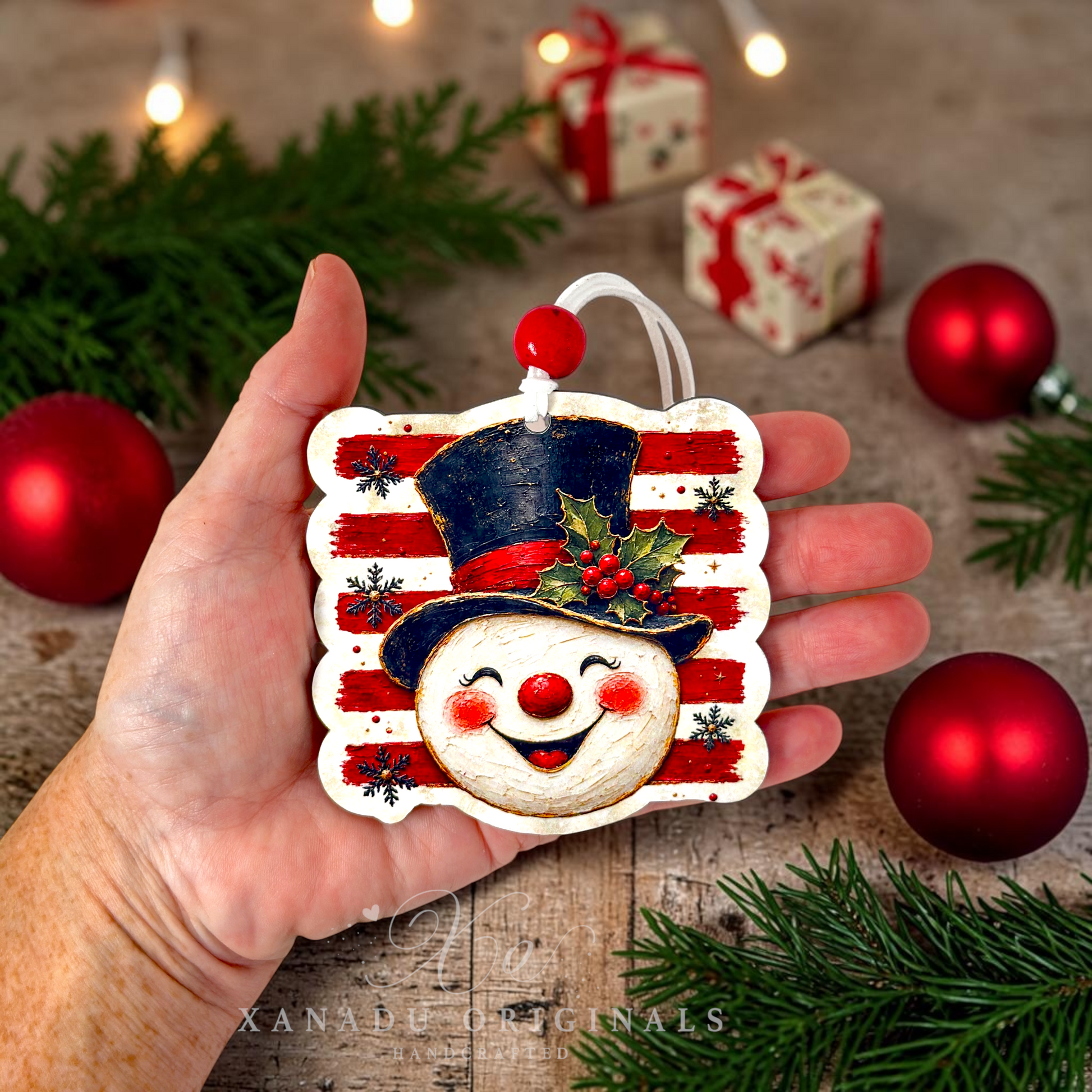 A hand holds a hand-painted wooden Christmas ornament shaped like a snowman wearing a navy blue top hat with red and white stripes, surrounded by festive holiday decorations including pine branches, red ornaments, and wrapped gift boxes in the background.