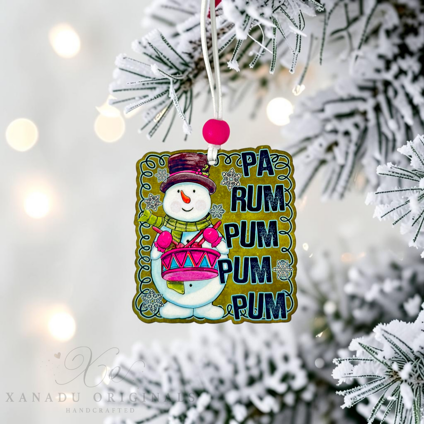 A handcrafted Christmas ornament featuring a cartoon snowman playing a pink drum, with the text 'PA RUM PUM PUM PUM PUM' in dark blue. The ornament hangs from a white string with a pink pom-pom, displayed against a blurred flocked Christmas tree with warm bokeh lights.