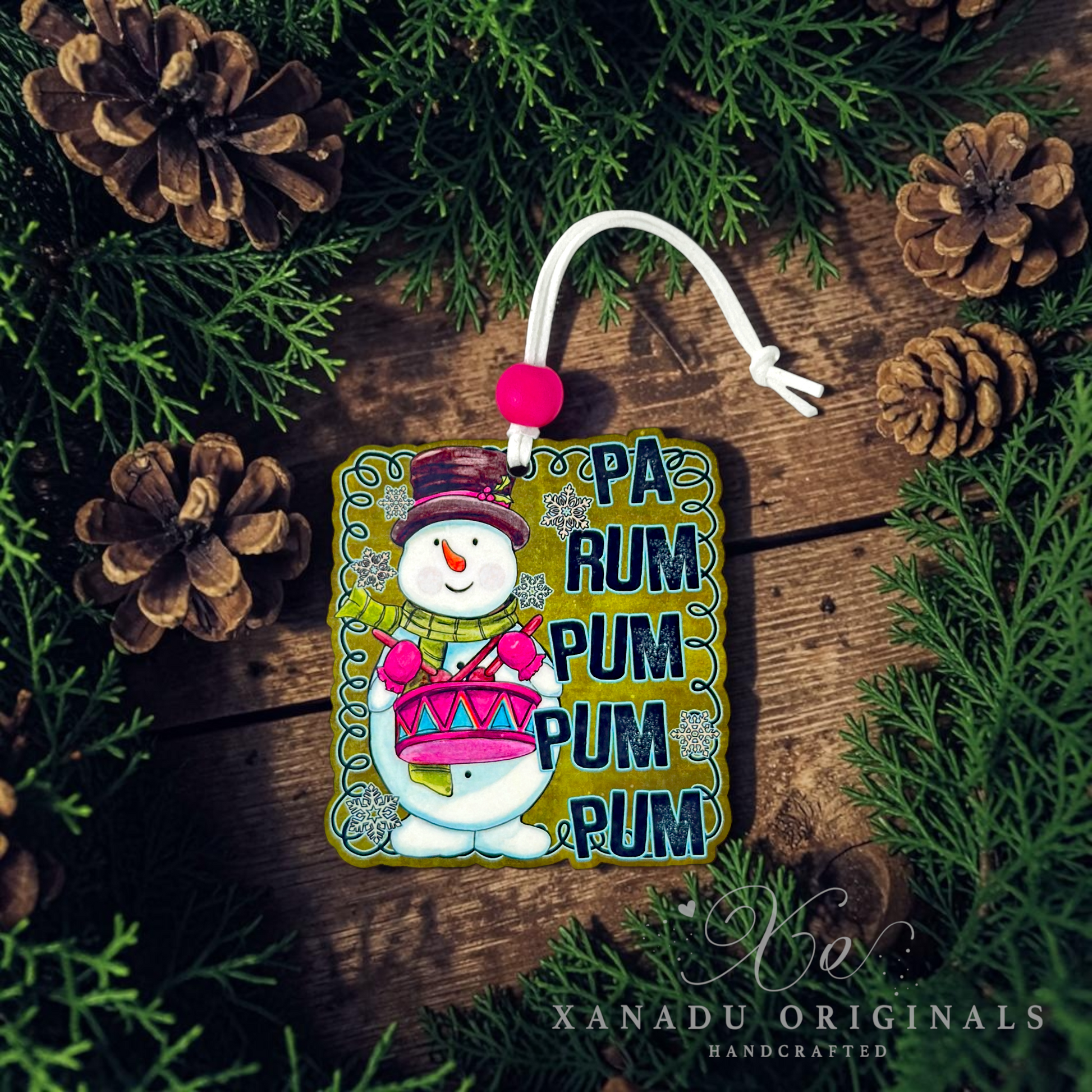 A square handmade ornament featuring a snowman playing a pink drum next to the text 'PA RUM PUM PUM PUM' in blue letters. The ornament is styled on a rustic wooden surface surrounded by pine branches and pinecones, with a white hanging loop and pink pom-pom.