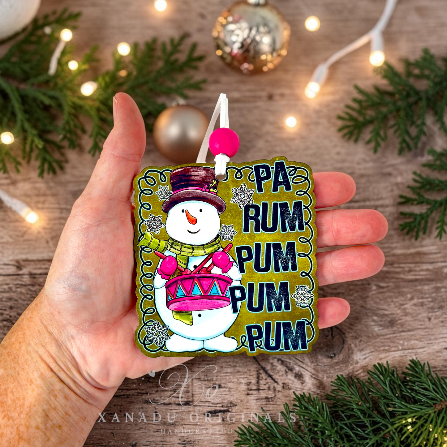 A hand holds a square wooden Christmas ornament featuring a snowman playing a pink drum next to the text 'PA RUM PUM PUM PUM PUM'. The background is a rustic wooden surface decorated with pine branches, fairy lights, and holiday ornaments.