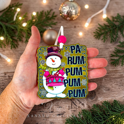 A hand holds a square wooden Christmas ornament featuring a snowman playing a pink drum next to the text 'PA RUM PUM PUM PUM PUM'. The background is a rustic wooden surface decorated with pine branches, fairy lights, and holiday ornaments.