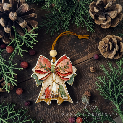 A decorative Christmas angel ornament with a large red bow, green pine branches, and a nativity scene, displayed on a dark wooden surface surrounded by pinecones and evergreen branches. A watermark for 'Xanadu Originals' is visible in the lower right corner.
