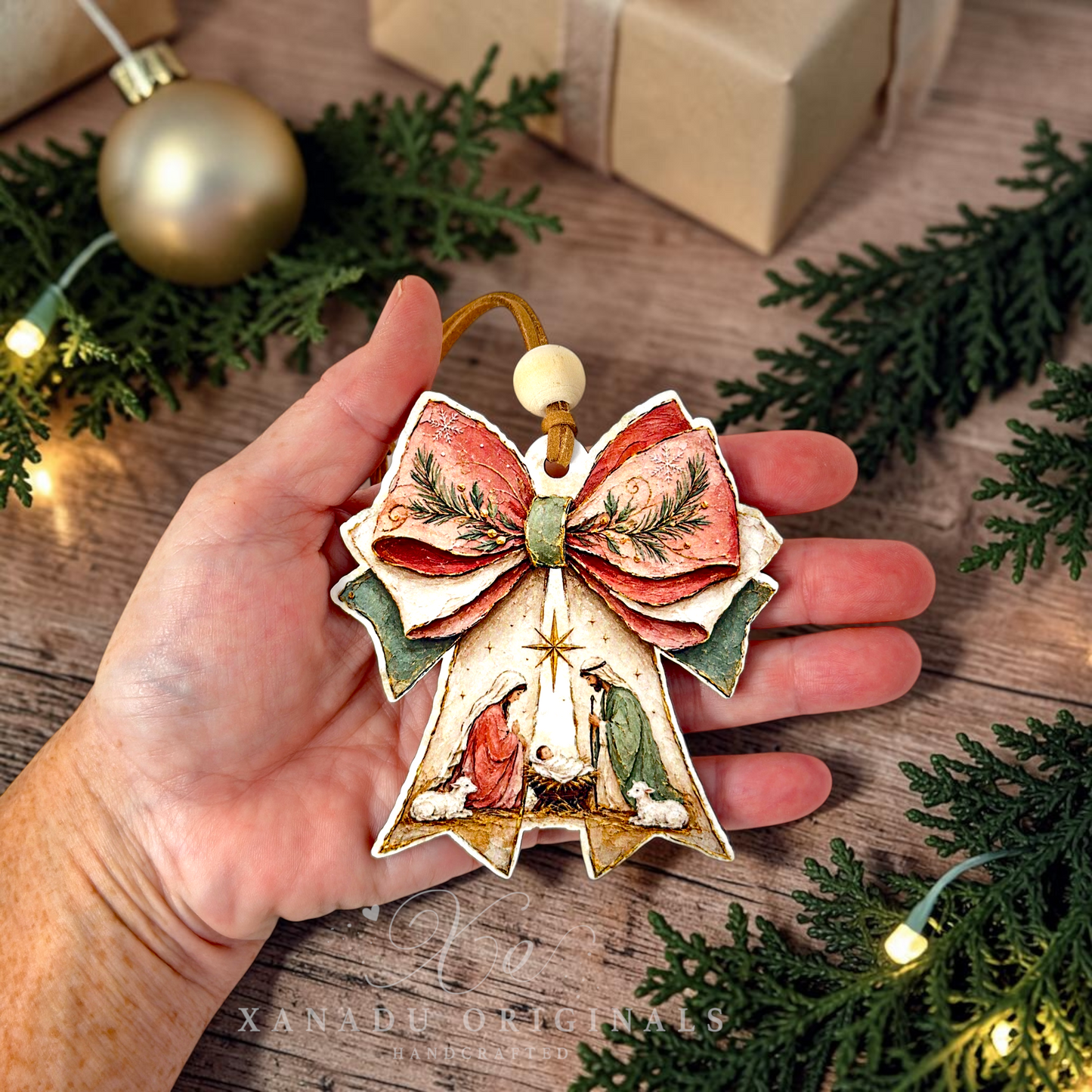 A hand holds a decorative Christmas angel ornament shaped like a bell with a large bow. The ornament has a cream base with red, green, and gold painted details, including a star and figures in robes.