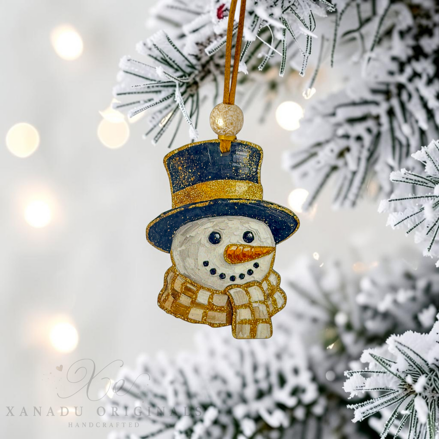 A handcrafted snowman Christmas ornament hanging from a gold string on a snowy flocked Christmas tree branch, with warm bokeh lights in the background. The ornament features a navy blue top hat with gold glitter trim, a cream-colored body, a checkered cream and gold scarf, and a carrot orange nose.