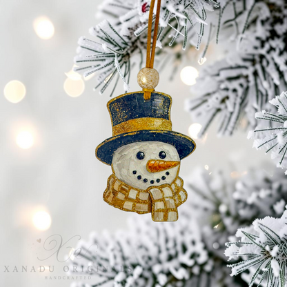 A handcrafted snowman Christmas ornament hanging from a gold string on a snowy flocked Christmas tree branch, with warm bokeh lights in the background. The ornament features a navy blue top hat with gold glitter trim, a cream-colored body, a checkered cream and gold scarf, and a carrot orange nose.