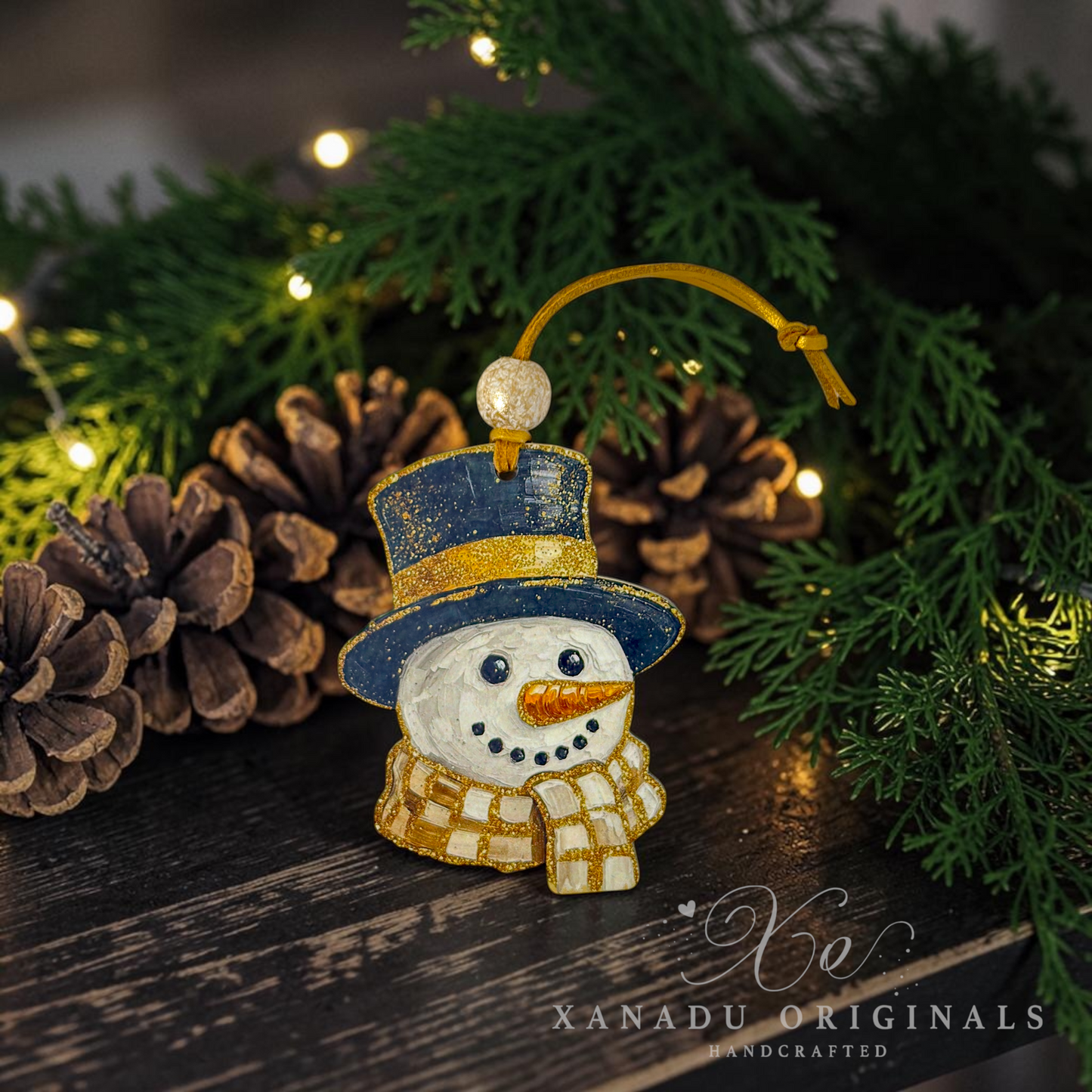 Handcrafted snowman Christmas ornament with a blue top hat, gold trim, white body, and gold plaid scarf, displayed on a dark wooden surface with pinecones and green pine branches under warm string lights. Xanadu Originals branding is visible in the lower right corner.