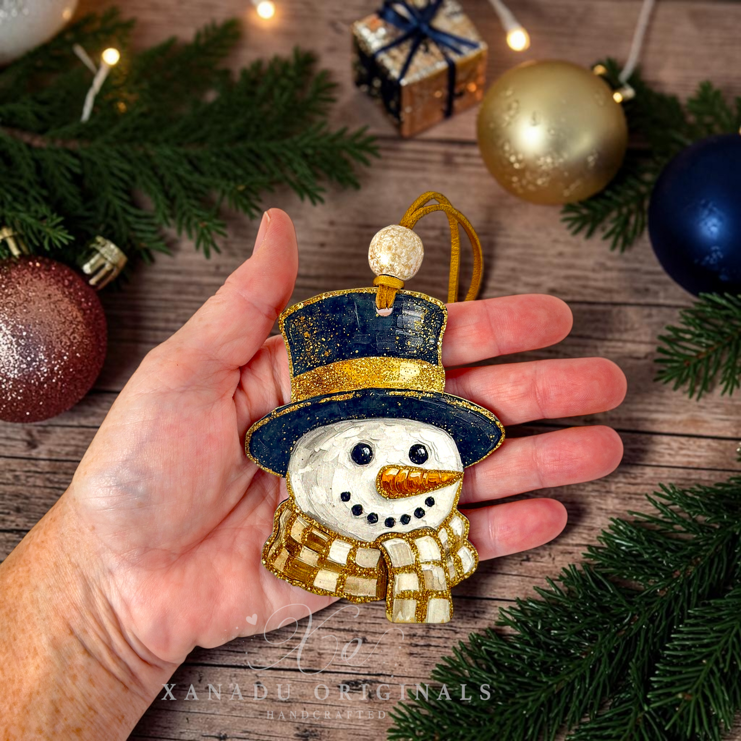 A hand holds a hand-painted snowman Christmas ornament with a navy blue top hat, gold trim, and a checkered scarf, surrounded by pine branches and other holiday ornaments on a wooden surface.