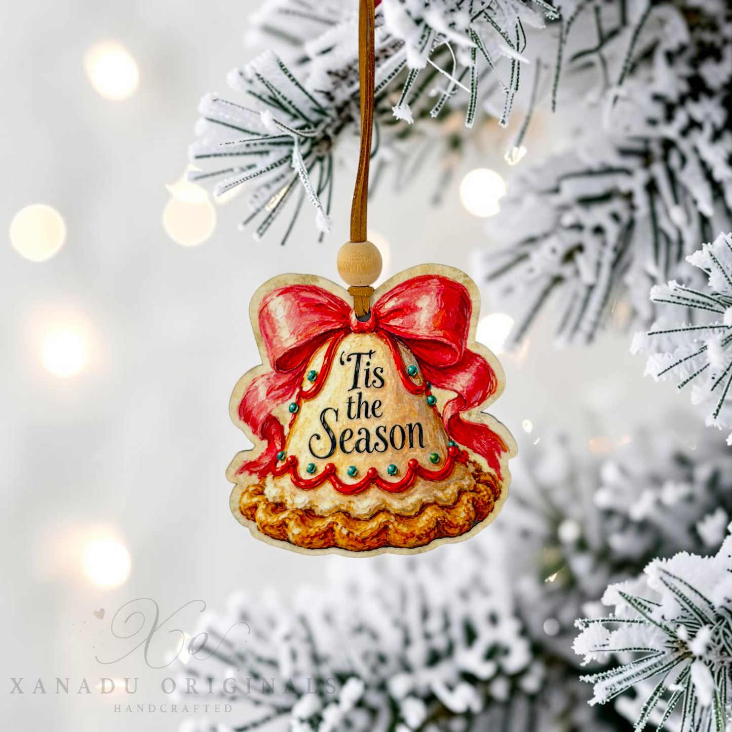 A handcrafted Christmas ornament shaped like a pie with a red bow and the words 'Tis the Season' in script, hanging from a flocked Christmas tree branch with warm bokeh lights in the background.