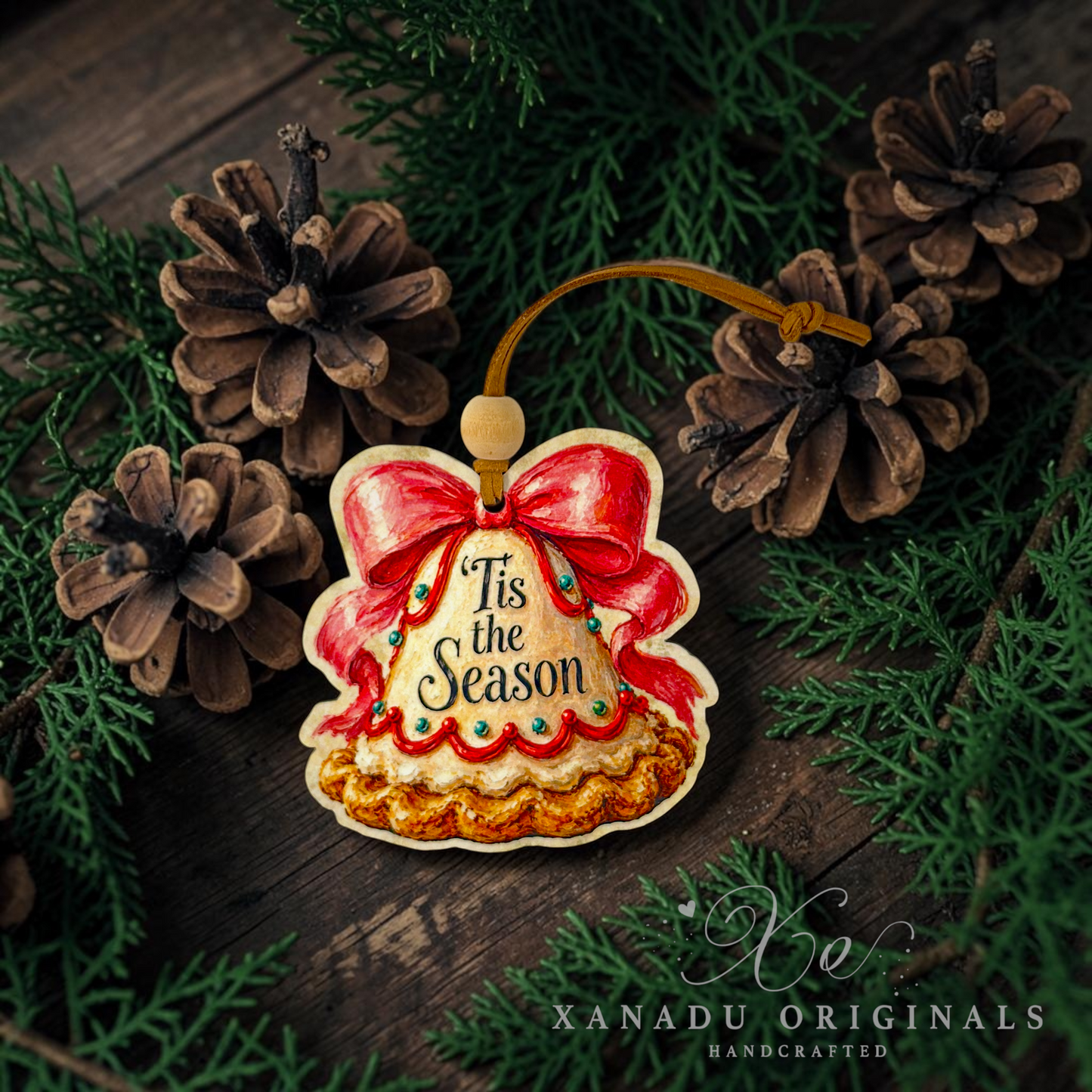 Handcrafted Christmas bell ornament with 'Tis the Season' text, decorated with red and gold icing and a wooden hanging loop, styled on a dark wood surface with pinecones and green pine branches. Xanadu Originals Handcrafted logo is visible in the bottom right corner.