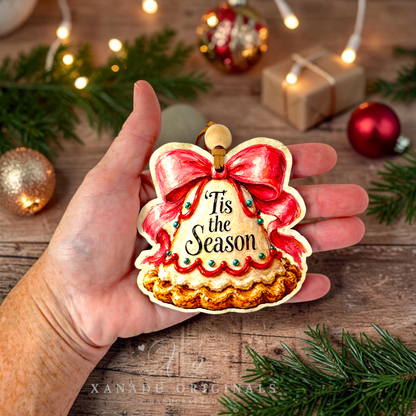 A hand holds a Christmas ornament shaped like a decorated bell, with the words “Tis the Season” in black script. The ornament has a red bow, red scalloped trim, and a gold-toned base.