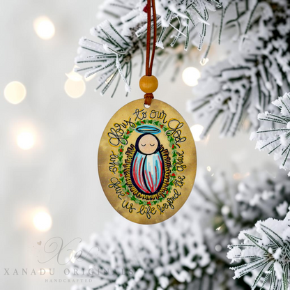 A round wooden Christmas ornament featuring a painted baby Jesus with a halo and the text 'Glor to Our God and Man Jesus Who Was Born to Us' hangs from a brown cord with a wooden bead, displayed on a snowy Christmas tree branch.