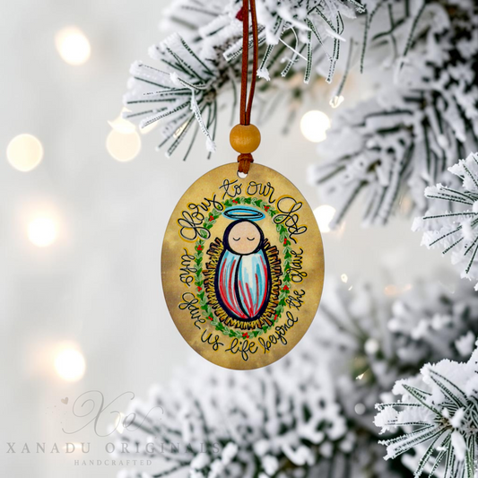 A round wooden Christmas ornament featuring a painted baby Jesus with a halo and the text 'Glor to Our God and Man Jesus Who Was Born to Us' hangs from a brown cord with a wooden bead, displayed on a snowy Christmas tree branch.