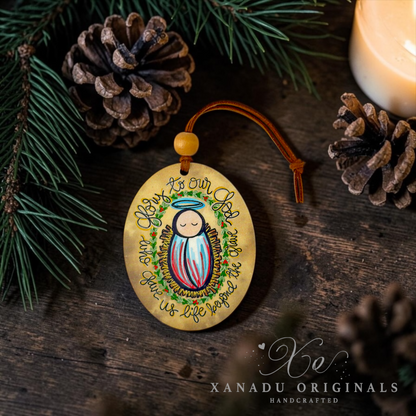 A round wooden Christmas ornament featuring a painted baby Jesus with the text 'Your is our God give us life through us life from you', displayed on a rustic dark wood background with pine needles, pinecones, and a lit candle. The brand 'XANADU ORIGINALS' is visible as a watermark in the bottom right corner.