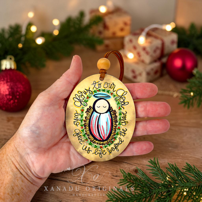 A hand holds a round wooden Christmas ornament featuring a painted baby Jesus with a halo and the text 'May Is Our Lord.