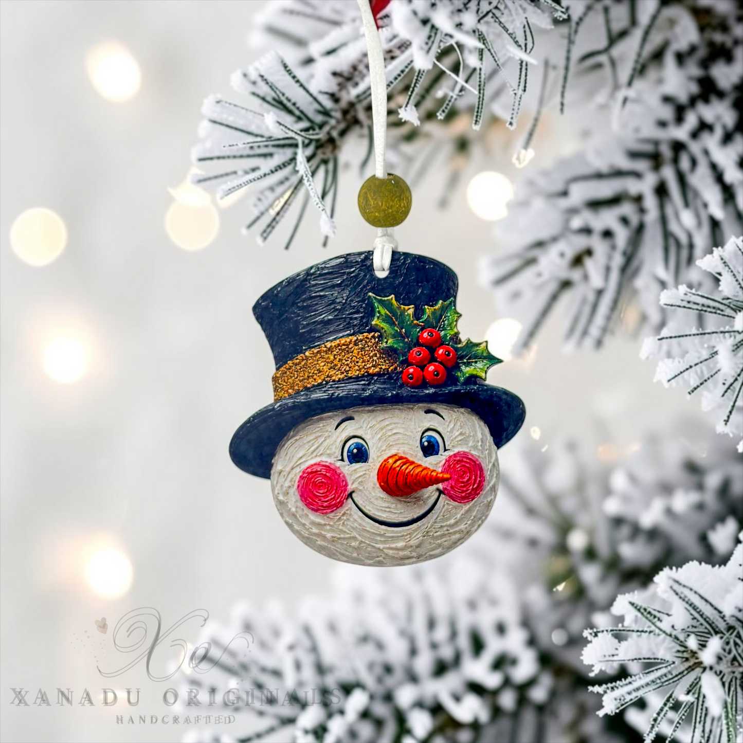 A handmade snowman Christmas ornament hanging from a snowy tree branch. The ornament features a round white face with blue eyes, a black top hat with a gold glitter band, green holly leaves, and red berries, and a carrot-shaped nose.