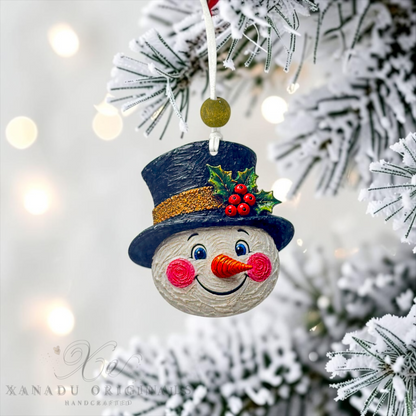 A handmade snowman Christmas ornament hanging from a snowy tree branch. The ornament features a round white face with blue eyes, a black top hat with a gold glitter band, green holly leaves, and red berries, and a carrot-shaped nose.