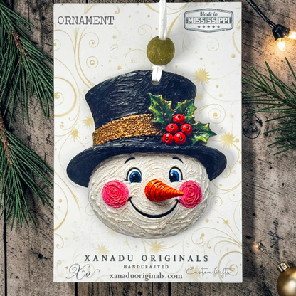 A handmade Christmas ornament card featuring a snowman face with a black top hat, gold glitter band, green holly leaves, and red berries. The card is displayed on a rustic wooden surface with pine branches and warm string lights.