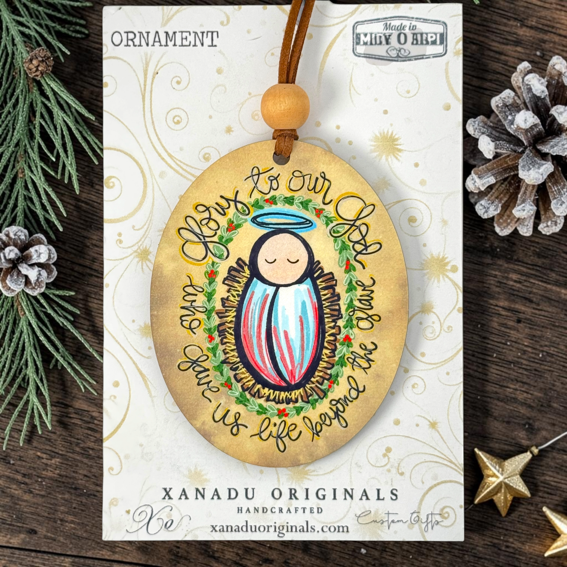 Handcrafted oval Christmas ornament featuring a baby Jesus illustration with a halo and the text 'Love to our God Your Life gives us life because the Christ', displayed on its branded card against a rustic pine and wood background.
