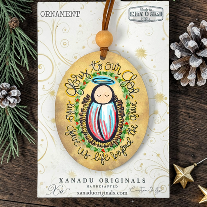 Handcrafted oval Christmas ornament featuring a baby Jesus illustration with a halo and the text 'Love to our God Your Life gives us life because the Christ', displayed on its branded card against a rustic pine and wood background.