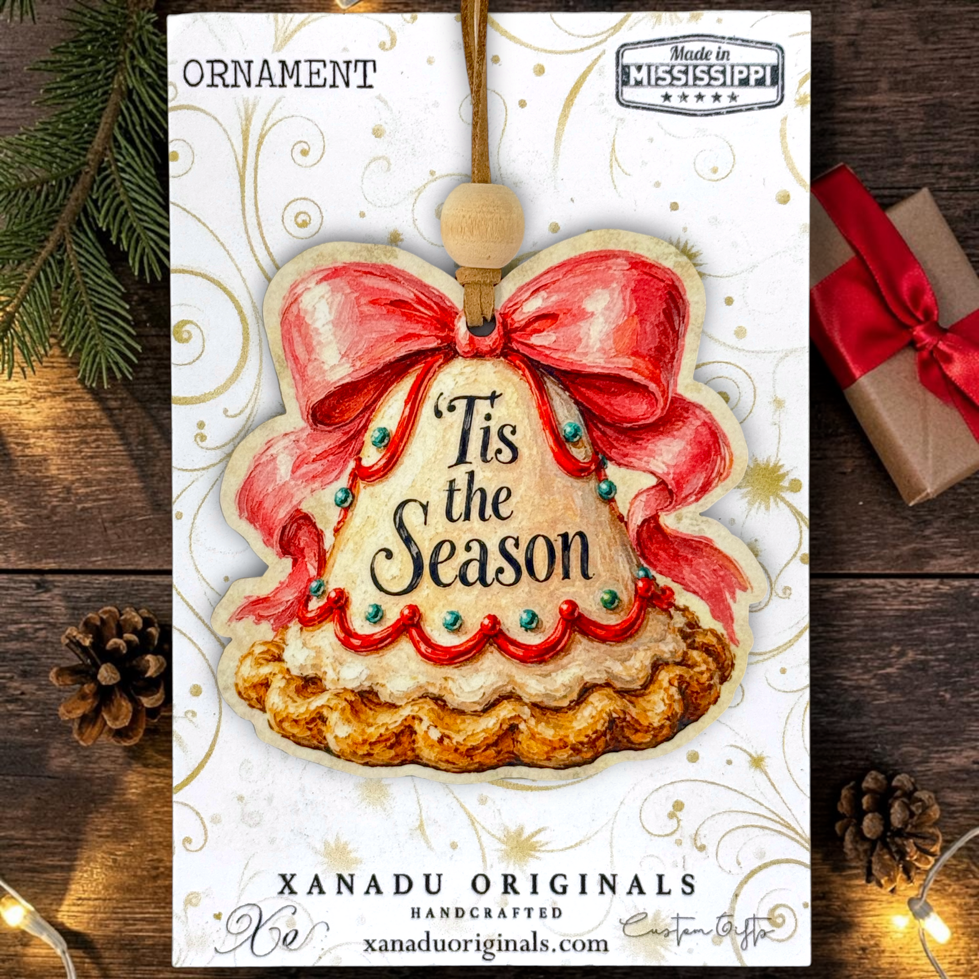 A Christmas ornament card or packaging design featuring a decorative ornament illustration with the words “Tis the Season,” surrounded by pine branches, a wrapped gift, and warm string lights on a wooden surface. Visible text includes “ORNAMENT,” “Made in Mississippi,” and “XANADU ORIGINALS.