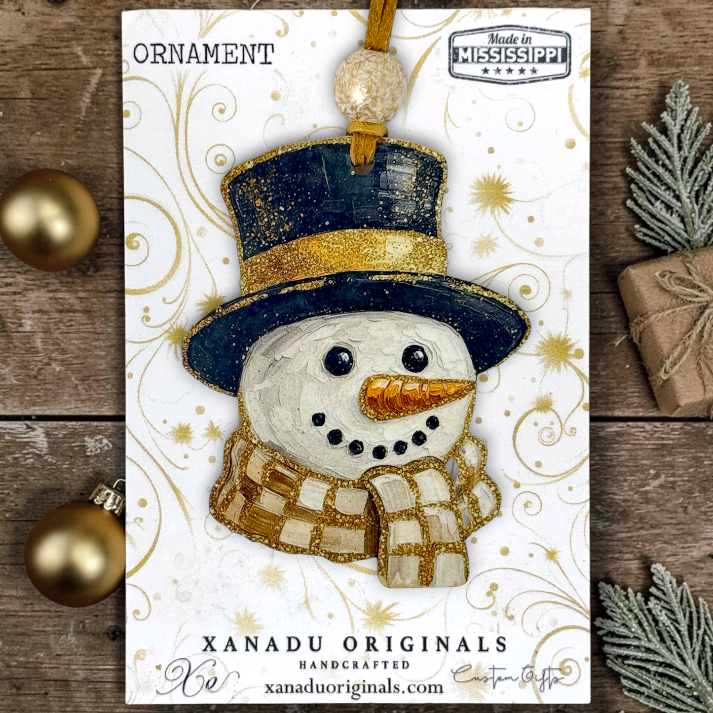 A handmade Christmas ornament card featuring a snowman ornament with a navy blue top hat, gold glitter band, white textured face, gold scarf, and gold beaded hanging loop. The card is displayed on a rustic wooden surface with holiday props including gold ornaments, evergreen branches, and a wrapped gift.