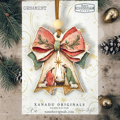 Handcrafted Christmas ornament card featuring a nativity scene ornament with a large red bow, green accents, and a wooden hanging loop, displayed on a rustic wood background with pine branches and gold ornaments.