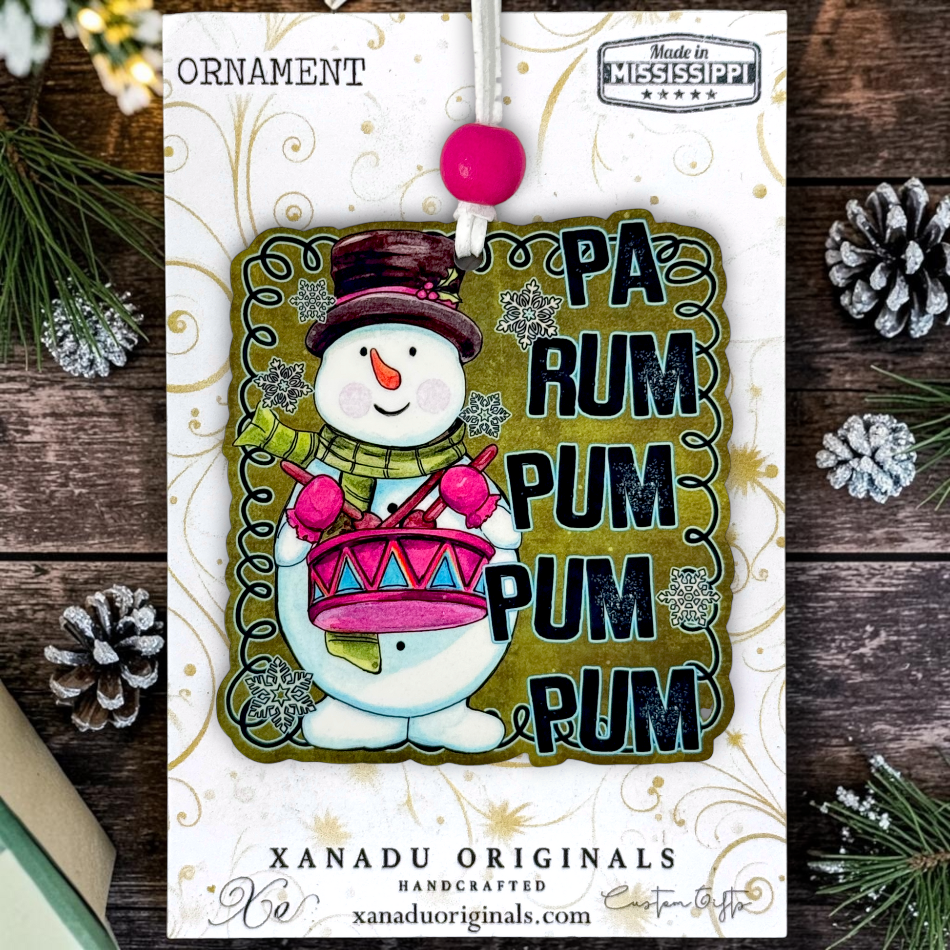A festive Christmas ornament card from Xanadu Originals featuring a snowman playing a pink and red drum, surrounded by the text 'PA RUM PUM PUM PUM'. The card is styled on a rustic dark wood background with pine needles and silver pinecones.