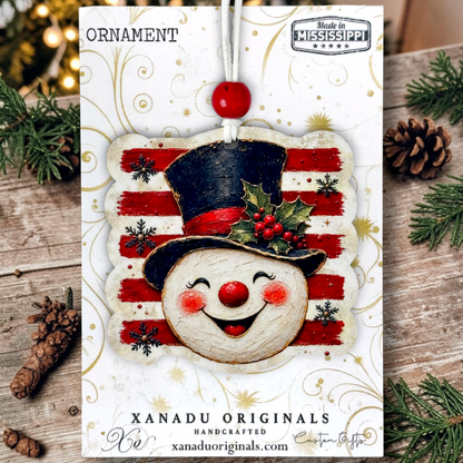 A festive Christmas ornament card featuring a handcrafted snowman ornament with a black top hat, red nose, red cheeks, and a holly accent on a red-and-white striped background. The card is displayed on a rustic wooden surface with pine branches, pinecones, and warm blurred lights around it.