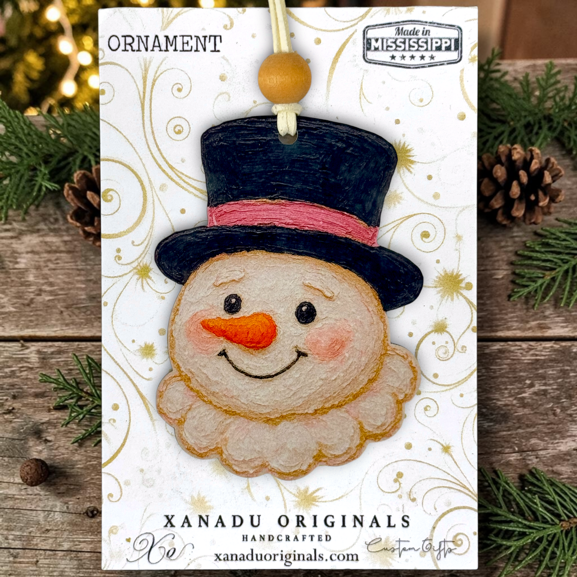 A snowman ornament carded on a branded Xanadu Originals display card, photographed on a rustic wood surface with pine branches and lights around it. The ornament has a black hat with a pink band, an orange carrot nose, and a cream beard.