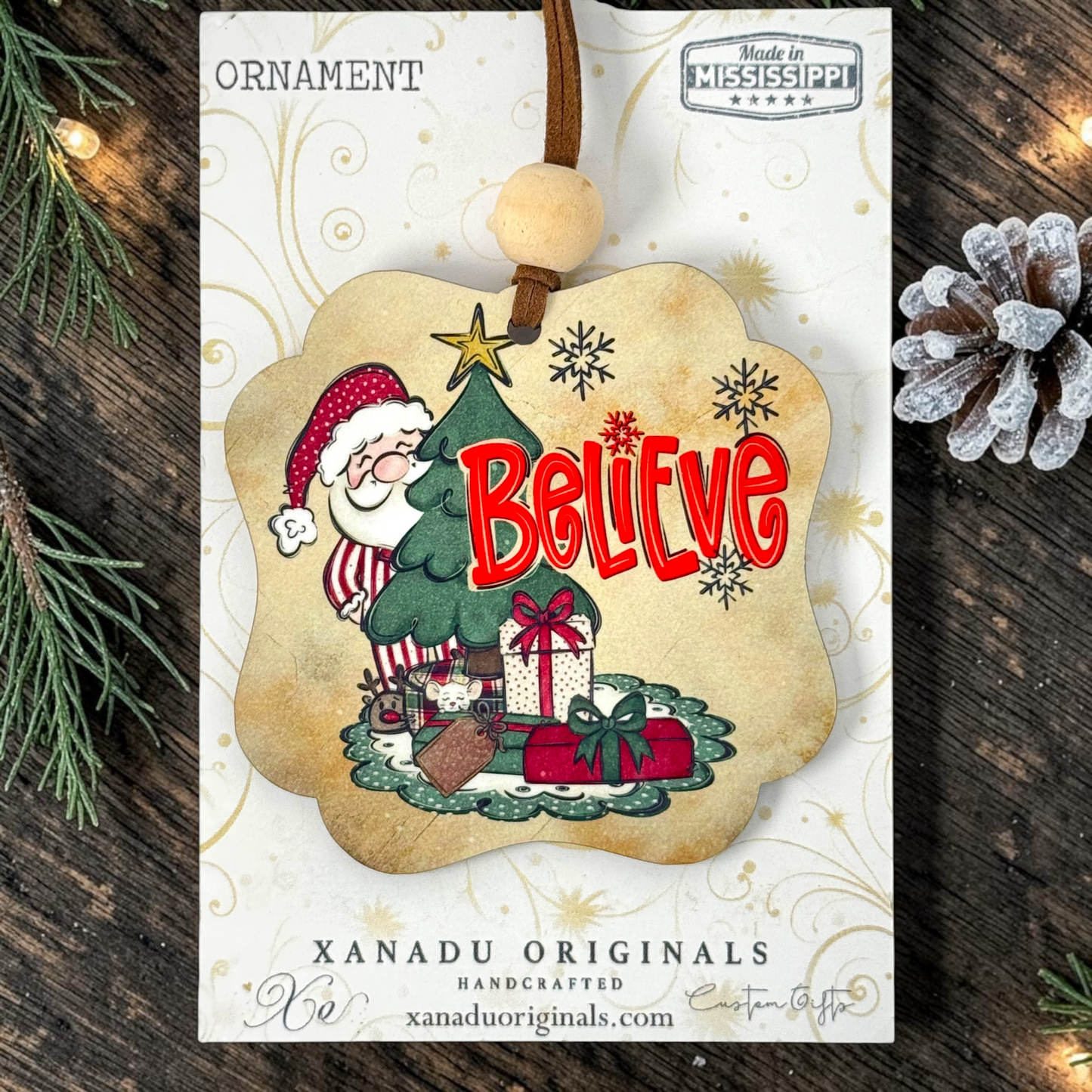 A Christmas ornament card featuring a handcrafted ornament design. The ornament has a scalloped beige border and shows Santa Claus sitting at a green Christmas tree with the word 'Believe' in red letters, surrounded by gifts and snowflakes.