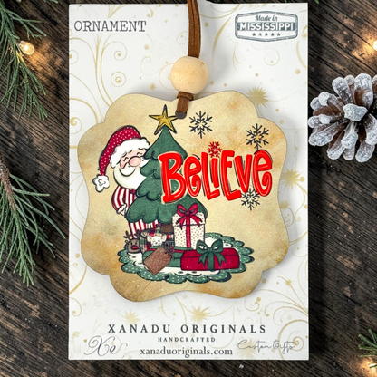 A Christmas ornament card featuring a handcrafted ornament design. The ornament has a scalloped beige border and shows Santa Claus sitting at a green Christmas tree with the word 'Believe' in red letters, surrounded by gifts and snowflakes.