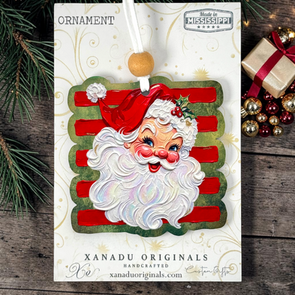 Handcrafted Christmas ornament by Xanadu Originals featuring a smiling Santa Claus face with white beard and red hat, set against red and green striped background details, hanging from a white ribbon with a wooden bead. The ornament is displayed on its branded card over a rustic wood background with pine branches and a wrapped gift.