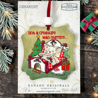 A Christmas ornament package from Xanadu Originals featuring a cute illustrated mouse in bed with a decorated tree and stockings. The ornament hangs from a red ribbon on a white backing card, styled with pine branches and small gift boxes on a dark wood surface.