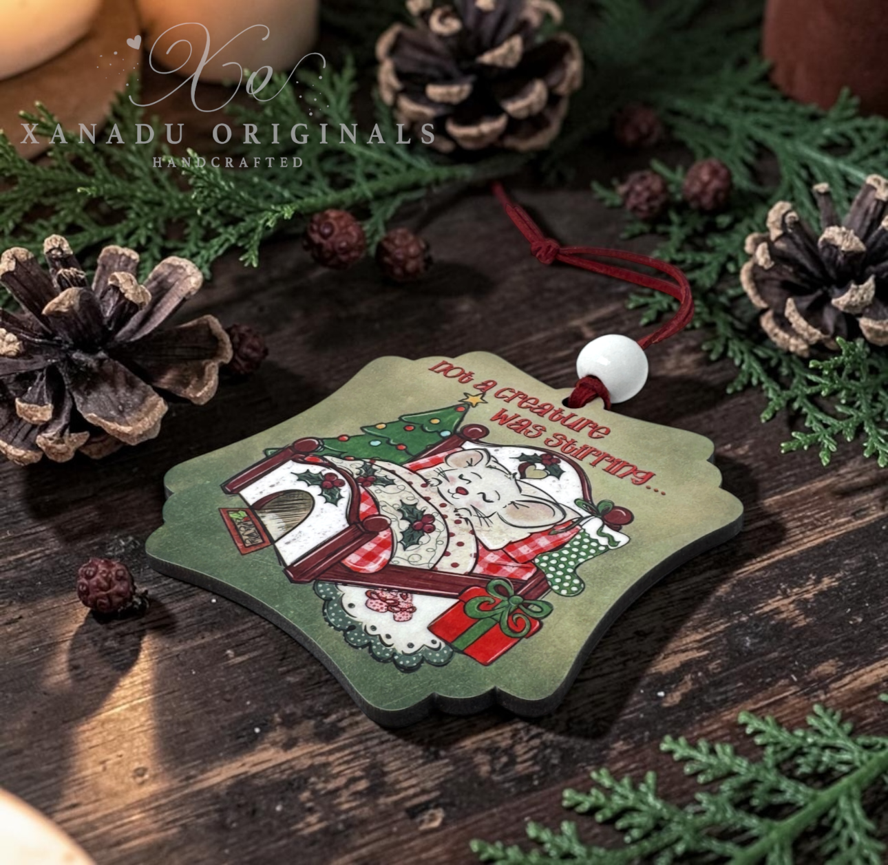 A decorative Christmas ornament shaped like a snowflake, featuring a cartoon mouse in a red plaid coat with a Christmas tree and presents, hanging from a red cord with a white bead. The ornament is styled on a dark wooden surface with pinecones and greenery.