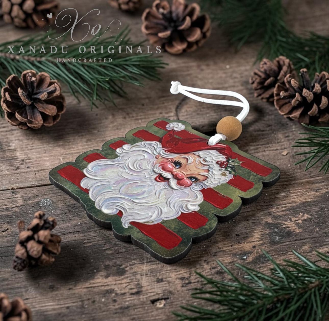 A handcrafted Christmas ornament shaped like a square sign with a painted Santa face, red and green striped background, and a white hanging cord, styled on a rustic wooden surface with pine branches and pinecones. The text 'XANADU ORIGINALS HANDCRAFTED' is visible in the upper left.