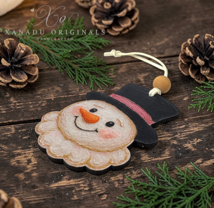 A handcrafted wooden snowman ornament by Xanadu Originals, featuring a painted face, black hat, pink band, and a small wooden bead on a cord, displayed on a rustic wooden surface with pine branches and pinecones.