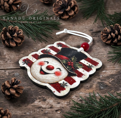 A handcrafted Christmas ornament shaped like a snowman face, featuring a distressed red and white striped pattern, a black hat with holly, and a white hanging cord with a red bead. The ornament is displayed on a rustic wooden surface surrounded by pine needles, pinecones, and a fir tree branch, with the brand name 'XANADU ORIGINALS' visible in the upper left.