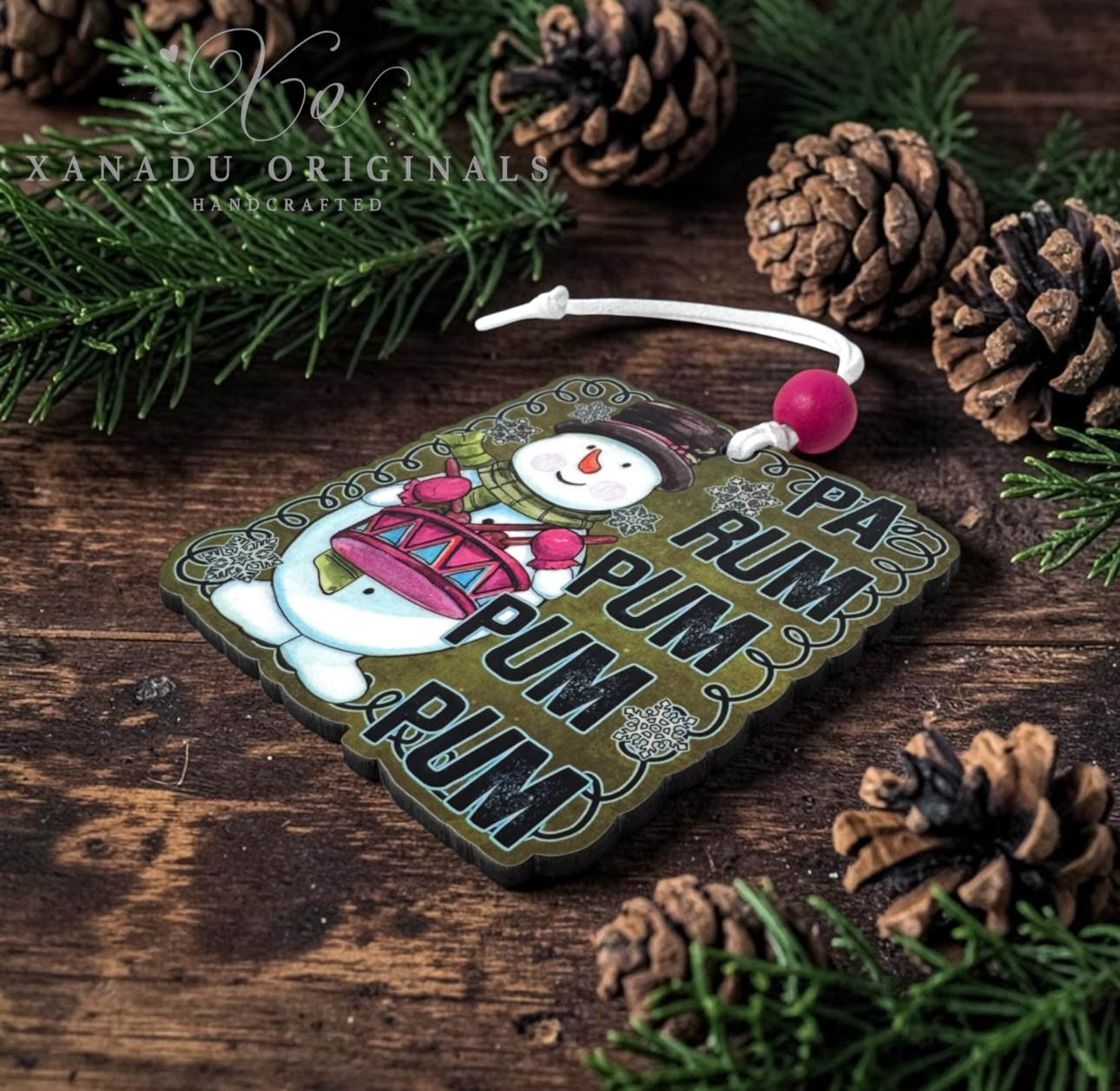 A square wooden Christmas ornament with a festive winter design, featuring a snowman holding a red gift box and the text 'PA-RUM PUM PUM' in large black letters. The ornament is decorated with a white cord and a pink bead, resting on a rustic dark wood surface surrounded by pine needles and pinecones.