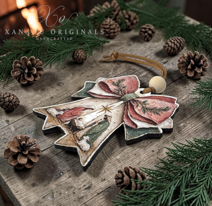 Handcrafted ceramic Christmas ornament shaped like a bow with a nativity scene, displayed on a rustic wooden surface surrounded by pine branches, pinecones, and a warm fire in the background. The brand watermark 'XANADU ORIGINALS' is visible in the top left corner.